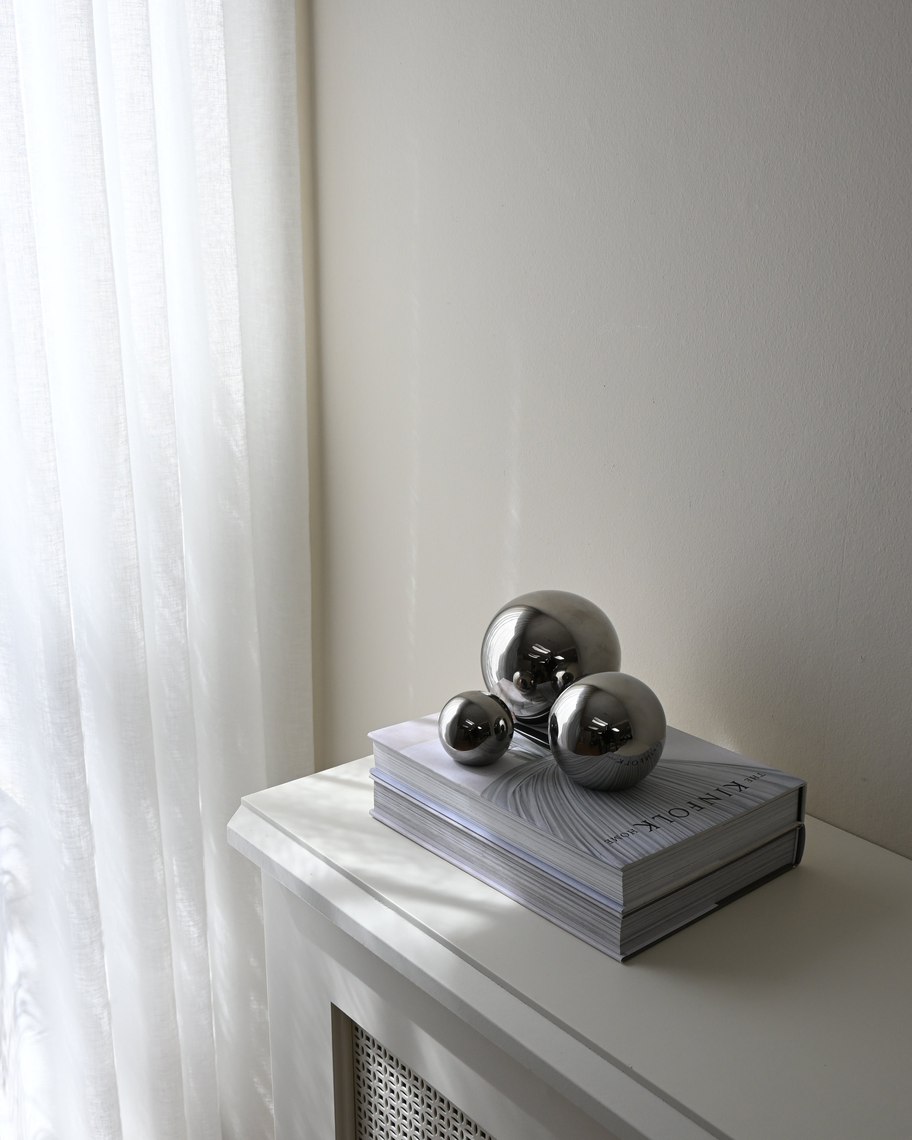 Trio of chrome spheres displayed on a book, placed on a white shelf near sheer curtains. Soft natural light creates a serene and minimalist atmosphere.