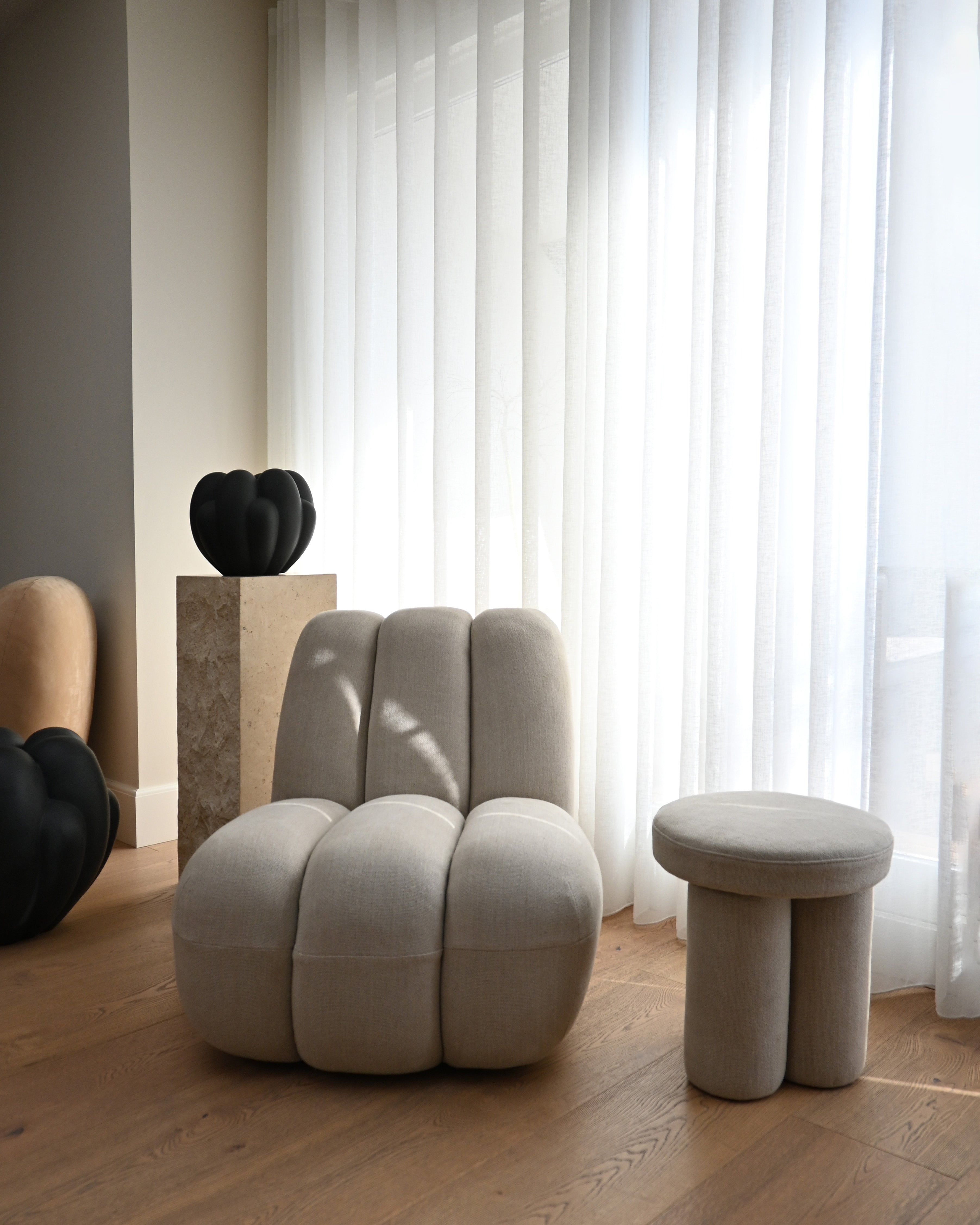 A serene living space showcasing a Sand-colored Toe Chair with thick, fluted upholstery and a matching Big Foot Stool. The room is filled with natural light coming through sheer white curtains. Decorative black objects rest on a travertine pedestal, while wooden flooring and minimalist décor enhance the space's calm, contemporary atmosphere