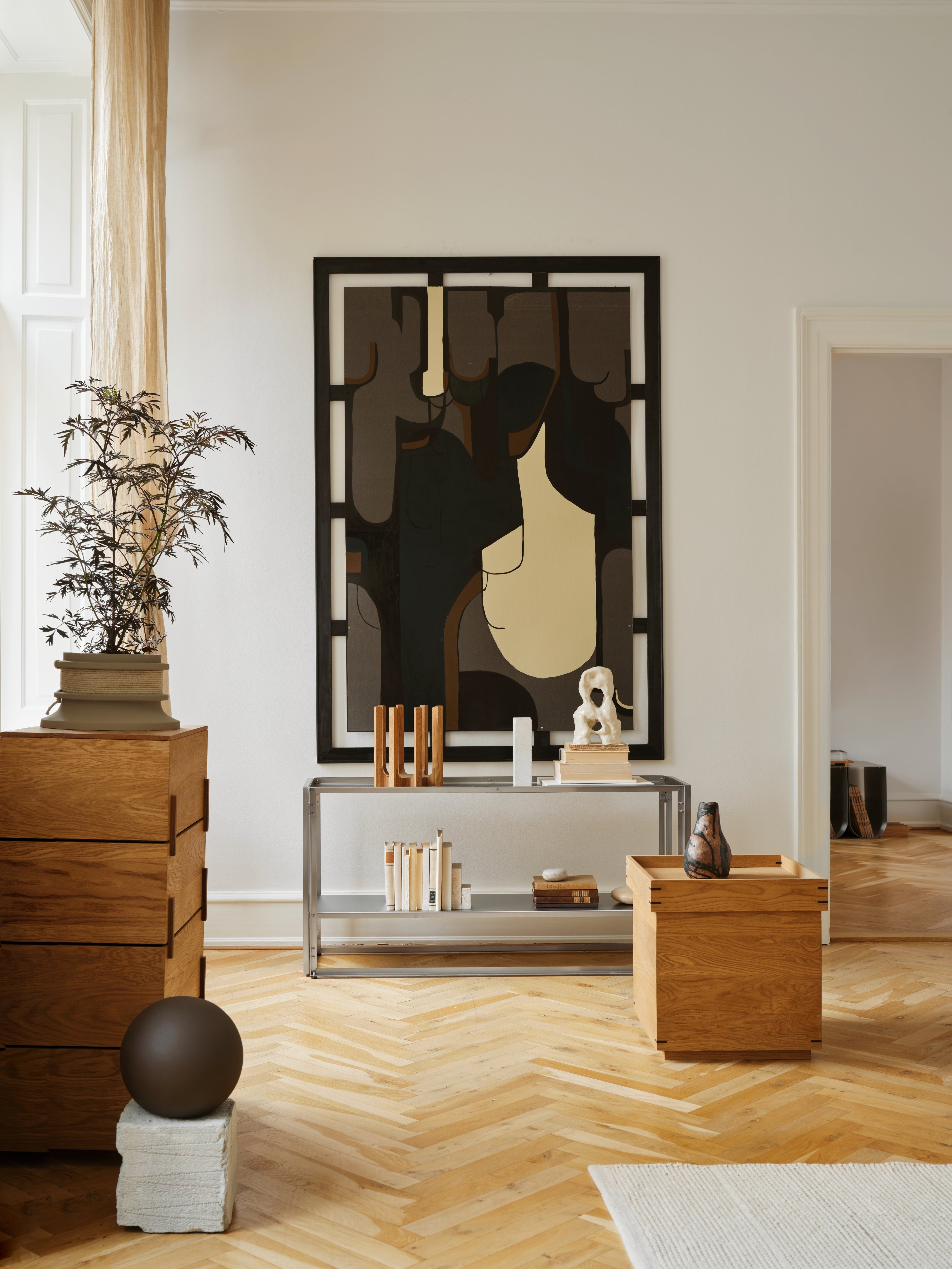 Modern interior with a stainless steel foldable console table against a wall, styled with books, sculptures, and decorative objects. A large abstract artwork hangs above, and the space features a wooden floor, oak furniture, and a potted plant.