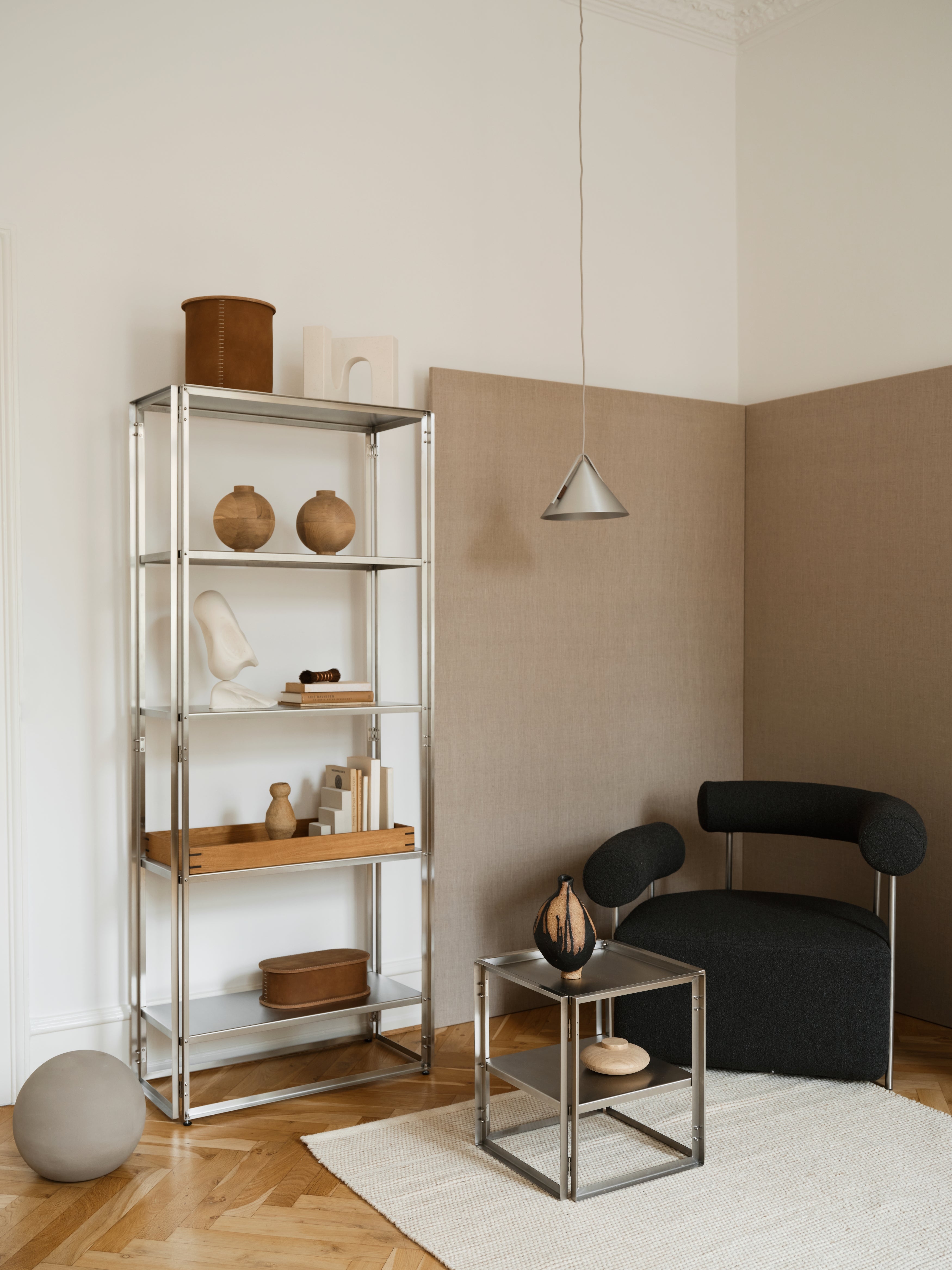 Modern interior featuring a stainless steel foldable shelf adorned with wooden vases, books, and sculptural décor. A black upholstered lounge chair sits beside a matching stainless steel side table with a ceramic vase, set against beige fabric-paneled walls and a white ceiling. The room has a minimalist, contemporary aesthetic.