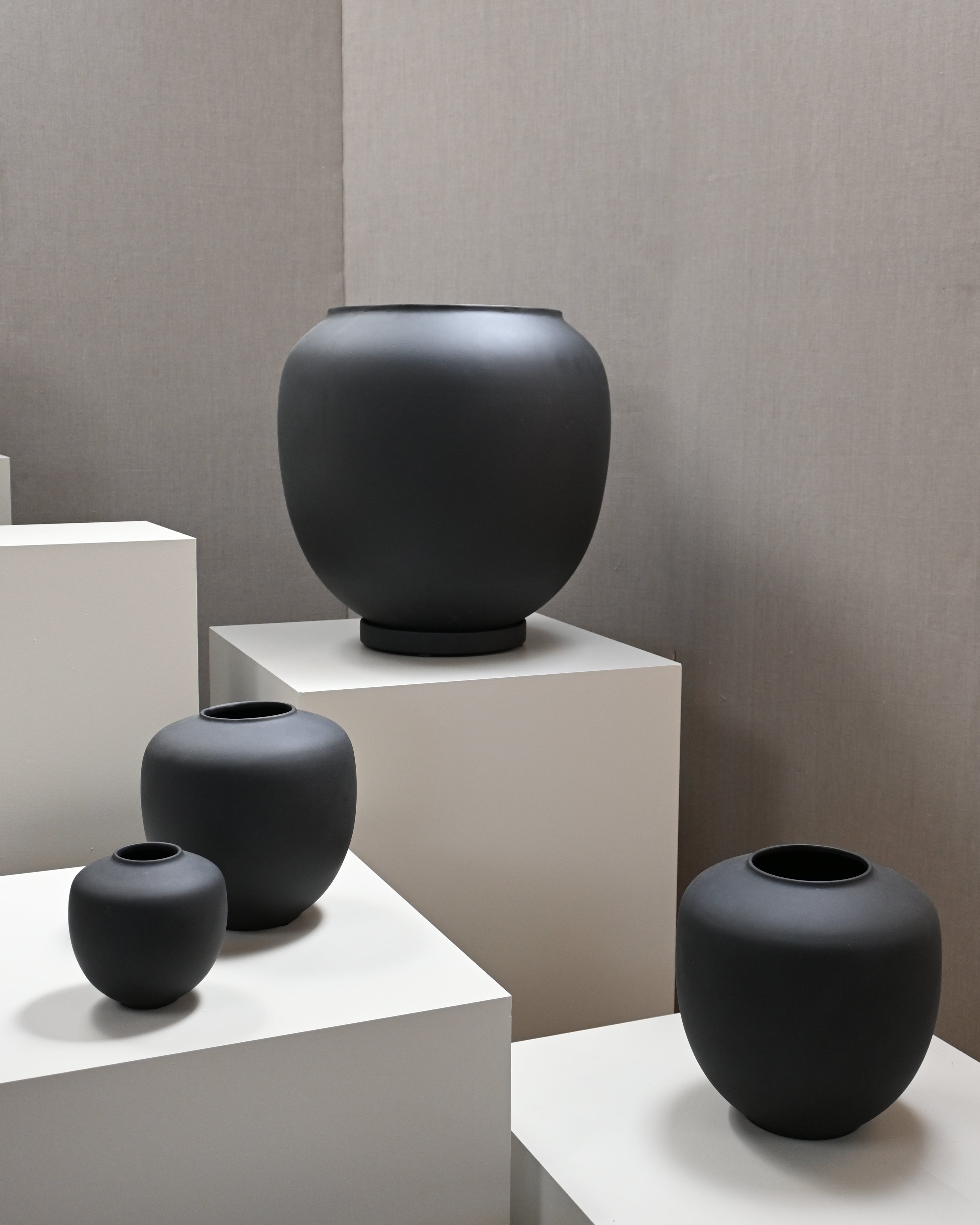 Collection of black matte ceramic vases of different sizes displayed on white pedestals in a minimalist setting.