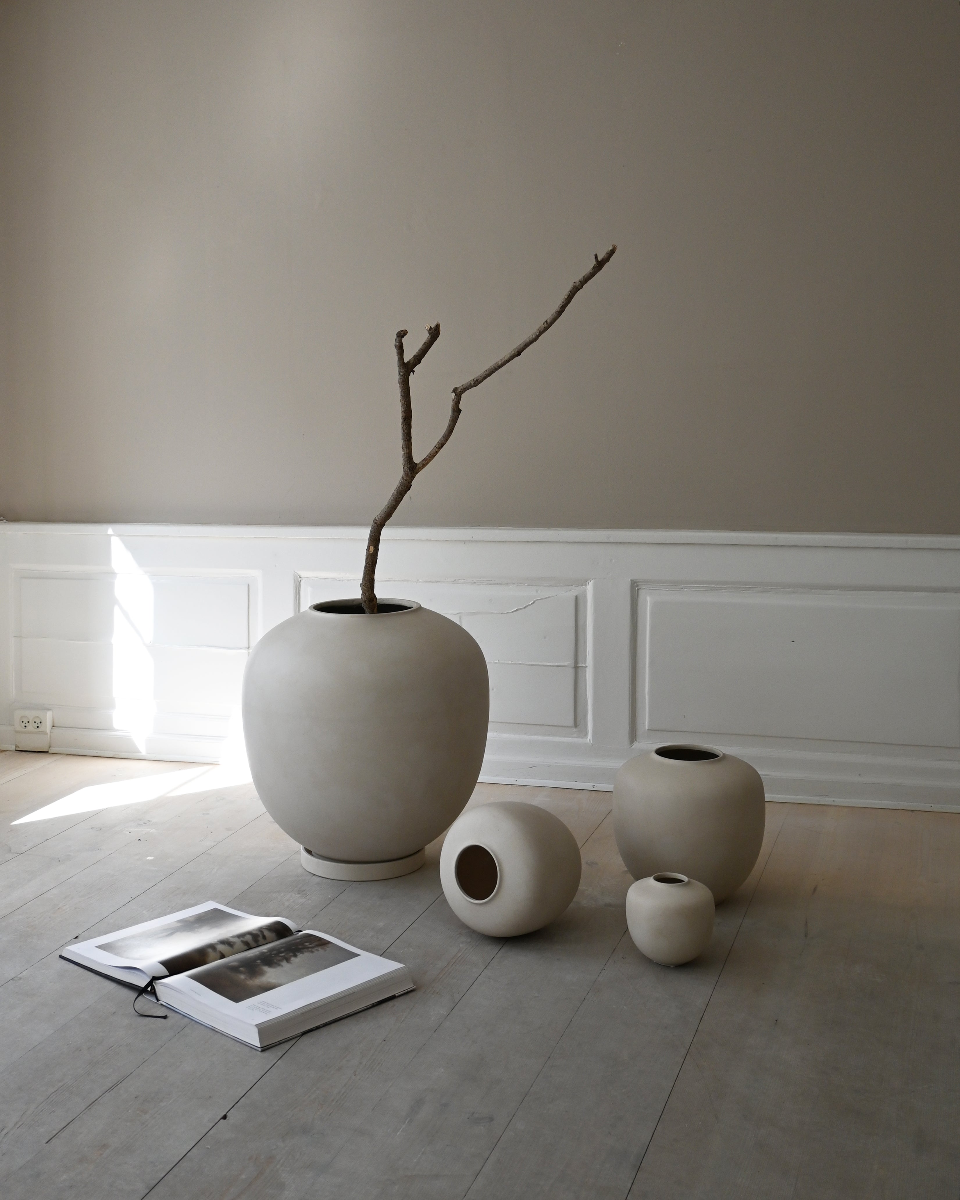 Group of beige ceramic vases of varying sizes, one holding a single branch, arranged on a wooden floor with an open design book beside them.