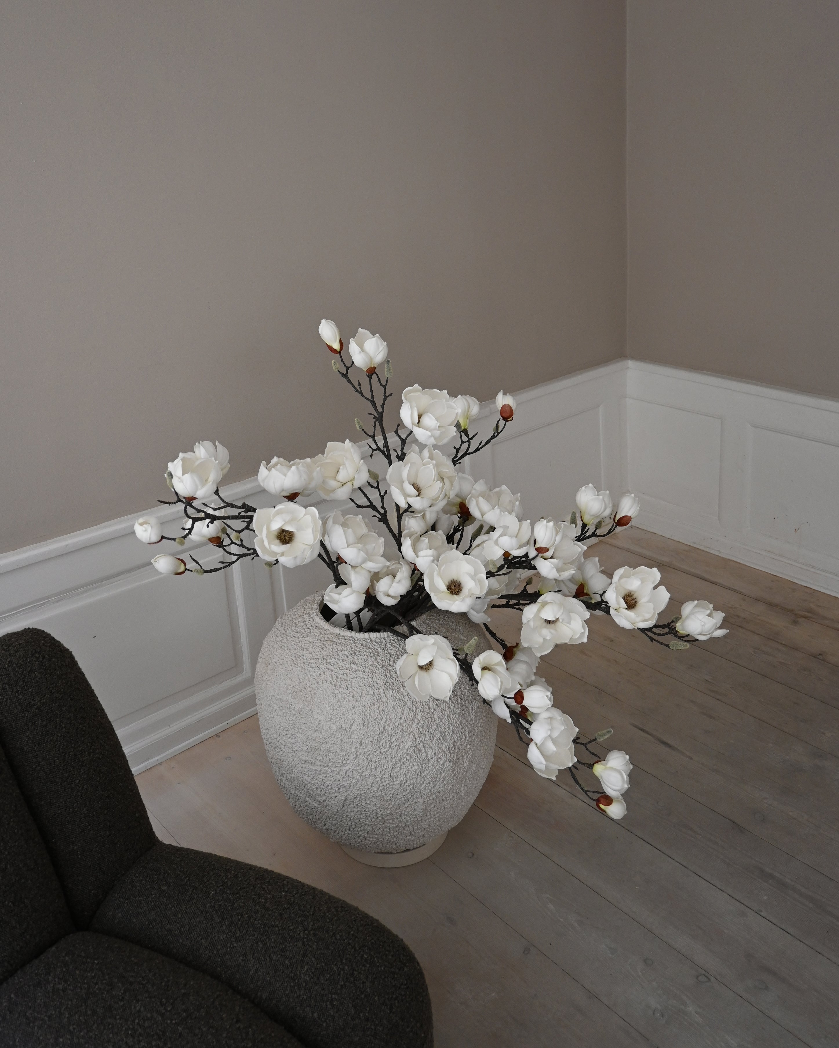 Large textured white vase with magnolia branches, placed beside a dark green chair in a softly lit room with white paneling