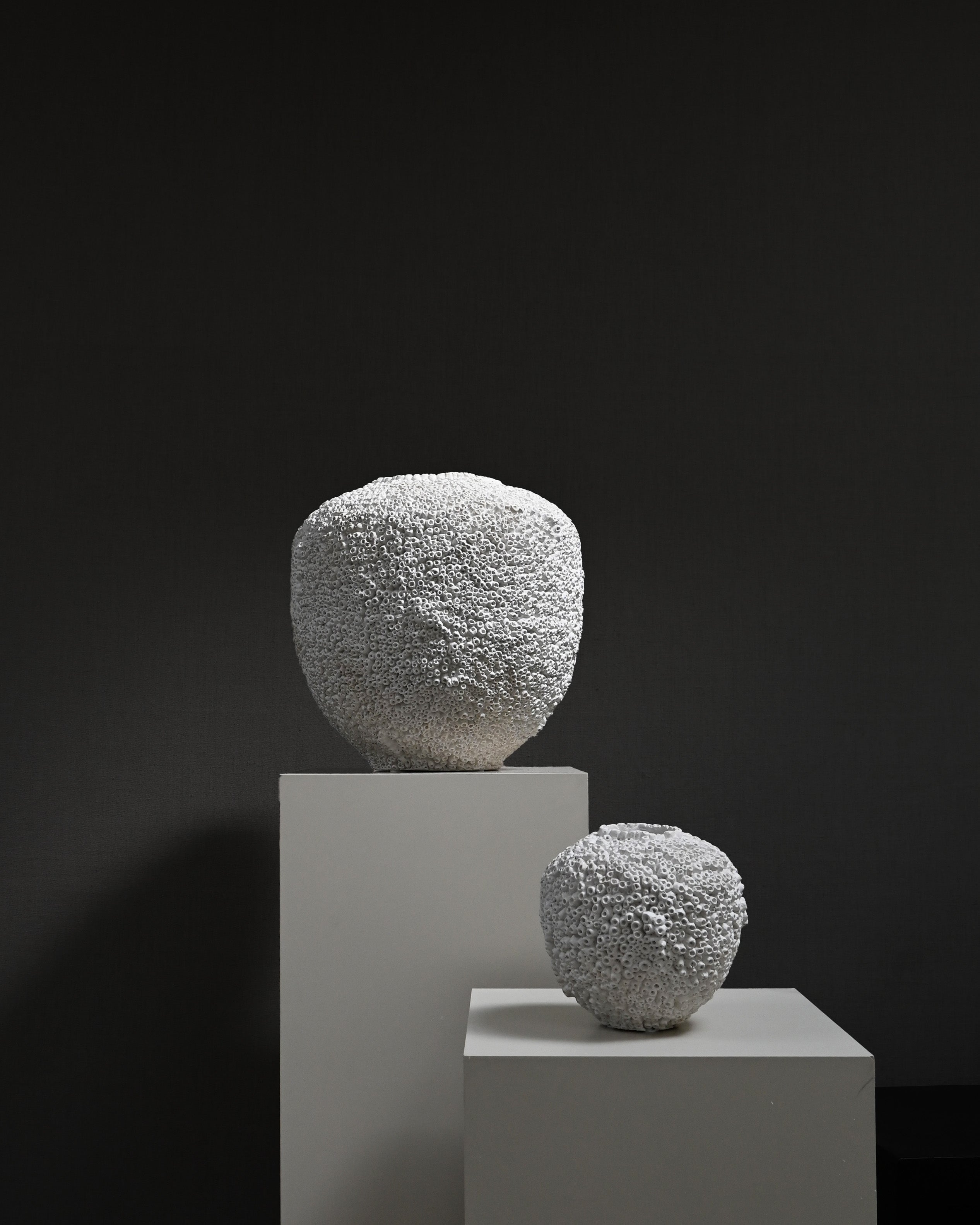 Two white, intricately textured ceramic vases displayed on white pedestals against a dark background.